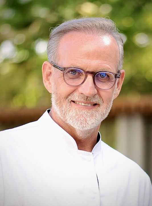 Retrato de un chef de mediana edad con barba canosa y gafas, sonriendo frente a un fondo natural, vestido con una chaqueta de cocina blanca.
