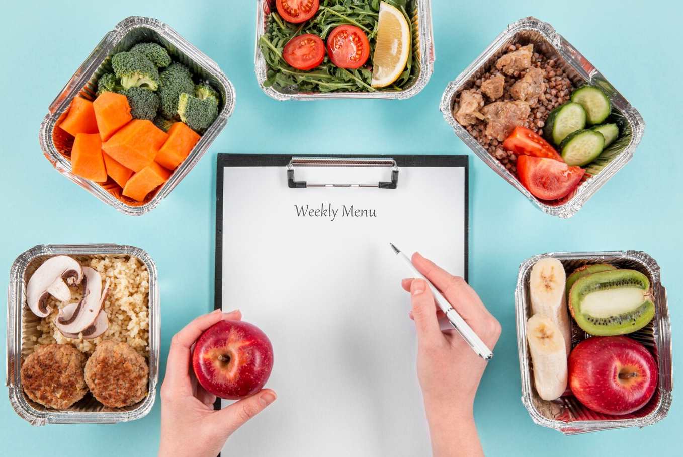 Persona escribiendo un menú semanal en una hoja, rodeada de recipientes de comida saludable con verduras, frutas y proteínas.