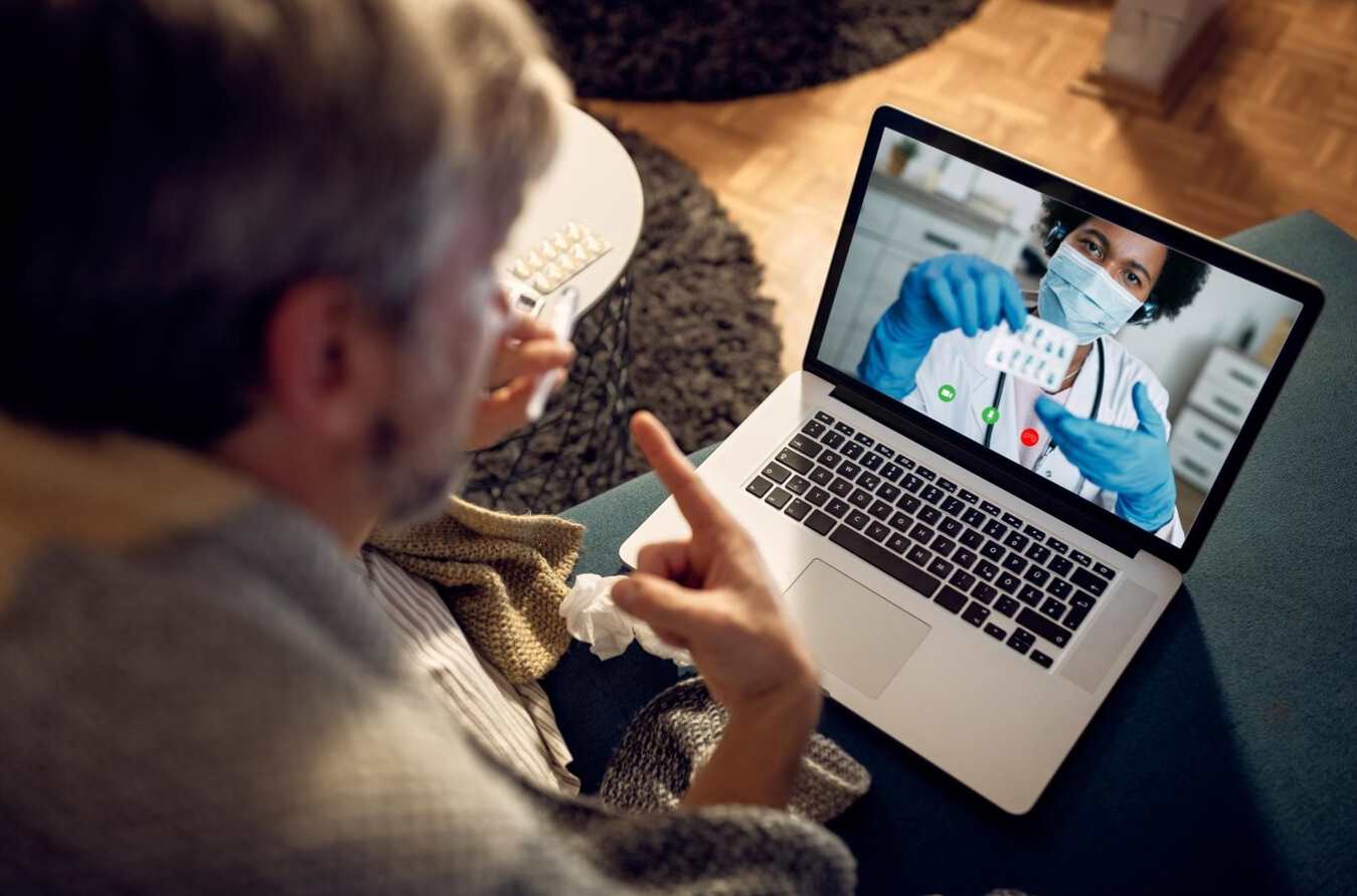 Hombre mayor participando en una consulta médica virtual a través de una computadora portátil, mientras un profesional de la salud le muestra información médica en pantalla.
